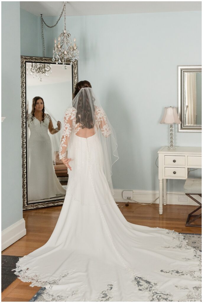 bride looking into mirror in bridal suite with dress train extended behind her and back towards camera at the manor house at prophecy creek