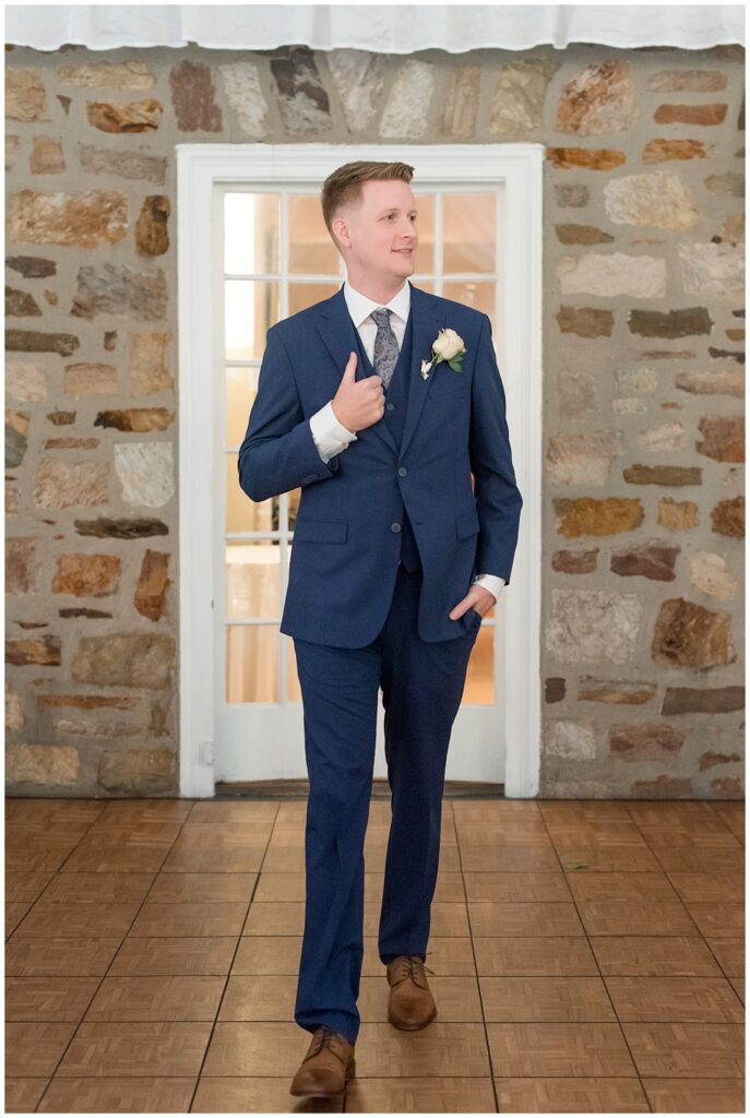 groom in navy blue suit holding edge of suit coat with right hand and looking left at the manor house at prophecy creek