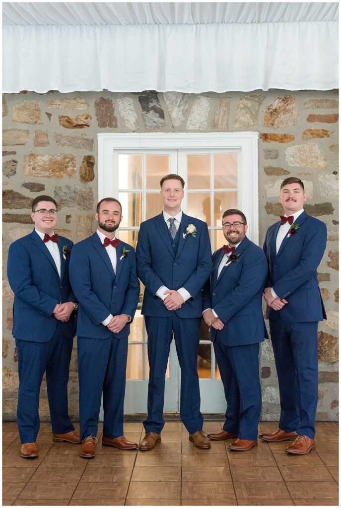 groom with his four groomsmen all wearing navy suits and smiling at camera by stone wall at the manor house at prophecy creek