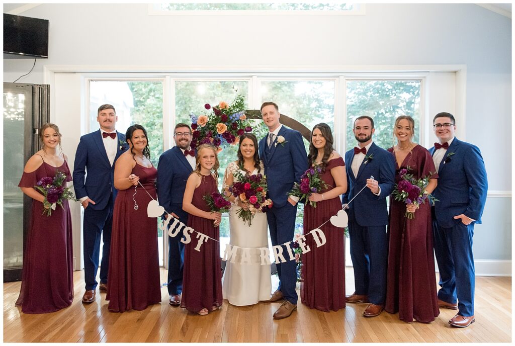 couple with their bridal party by wooden floral display as bridal party holds "just married" banner in front of couple in ambler pennsylvania