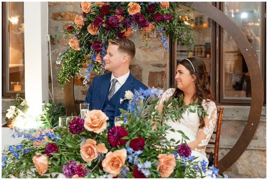 couple sitting and smiling during bridal party speeches by large colorful floral table display at the manor house at prophecy creek