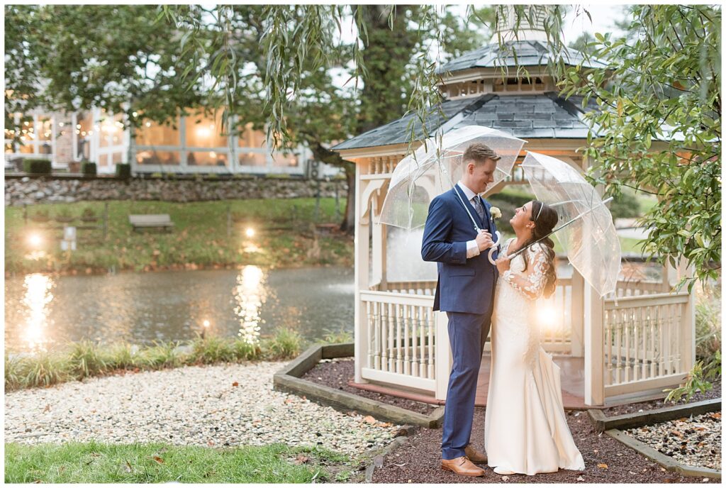couple holding clear umbrellas outdoors on rainy wedding day and smiling at each other by white gazebo at the manor house at prophecy creek