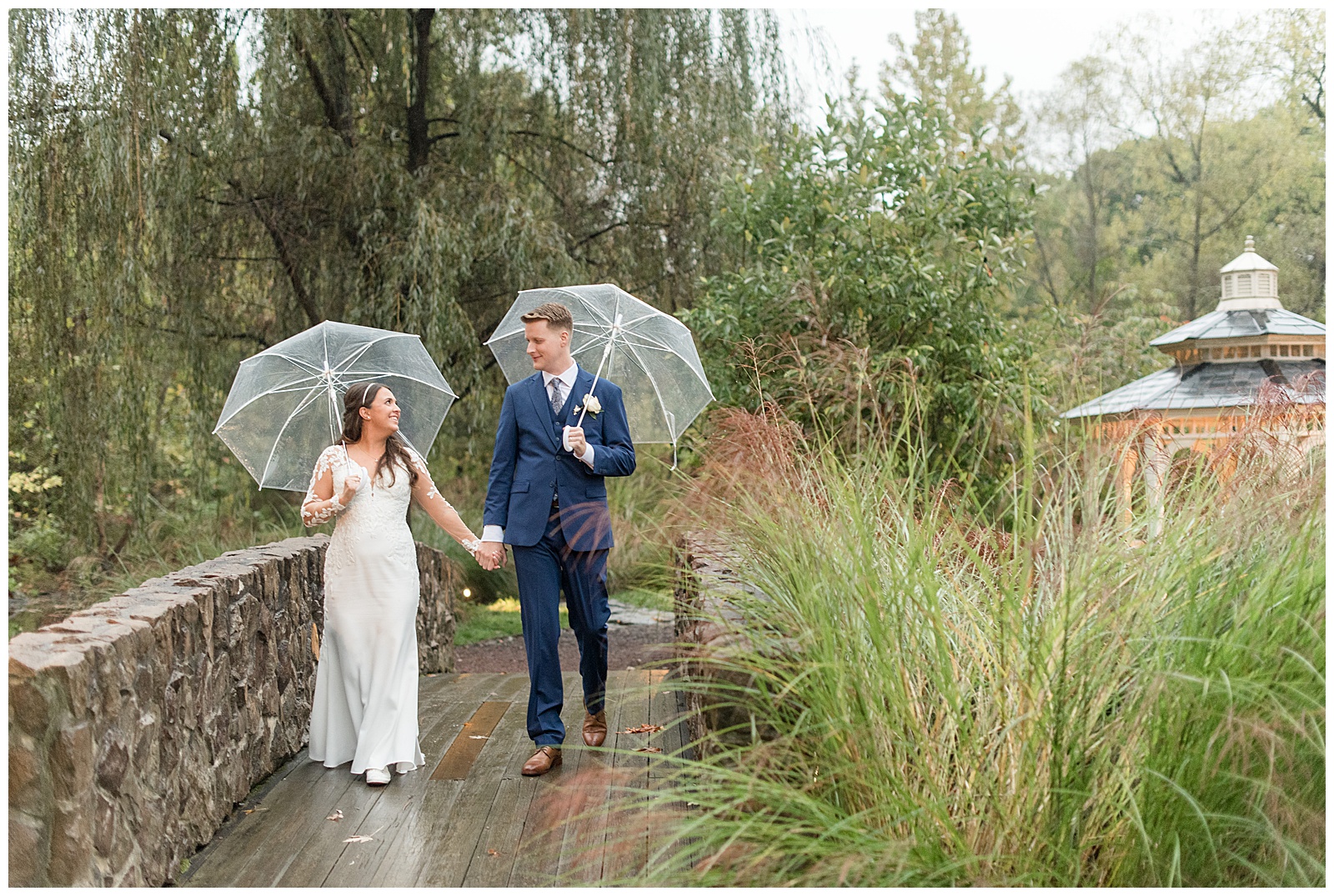 couple holding hands and walking along stone wall holding clear umbrellas at the manor house at prophecy creek