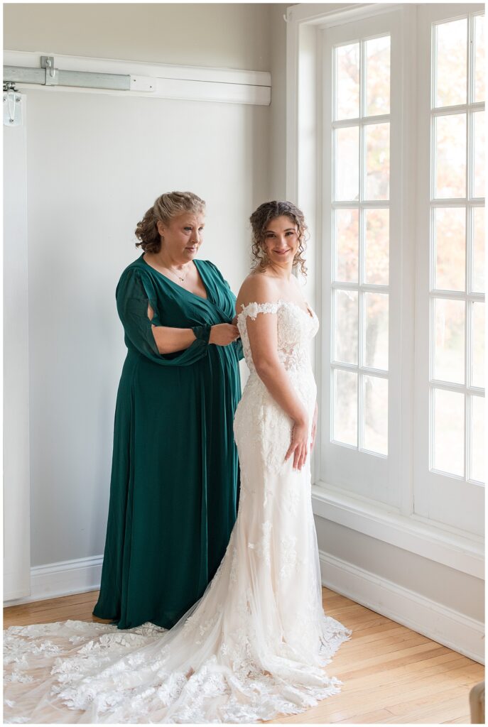 bride's mom in long dark green gown zipping the back of bride's lacy off-the-shoulder white wedding gown in bridal suite at lauxmont farms