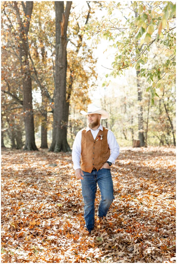 groom in white shirt with brown vest, blue jeans, and white cowboy hat looking to his right in forest of fallen leaves at lauxmont farms