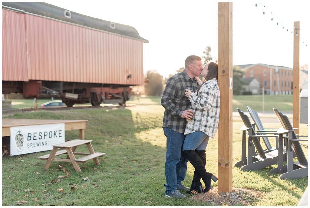woman leaning against wooden post as man kisses her with red train in background at bespoke brewery in strasburg pa