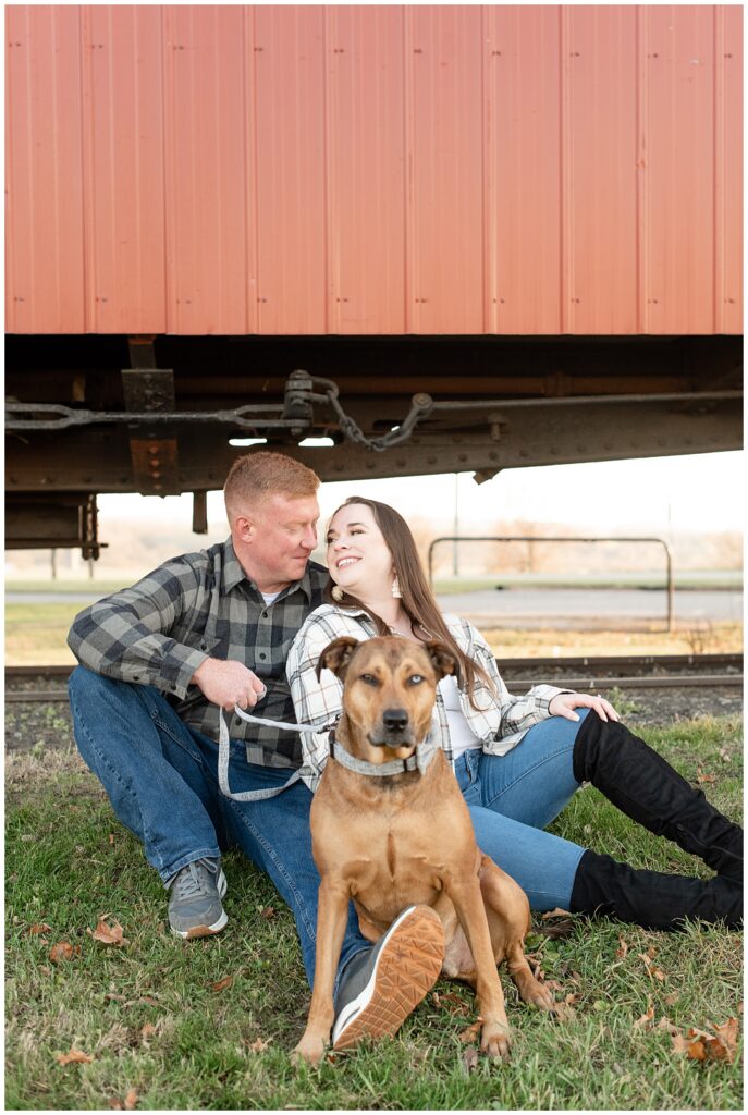couple sitting on the grass by red train with their pup looking at the camera in front of them at brewery in lancaster pennsylvania
