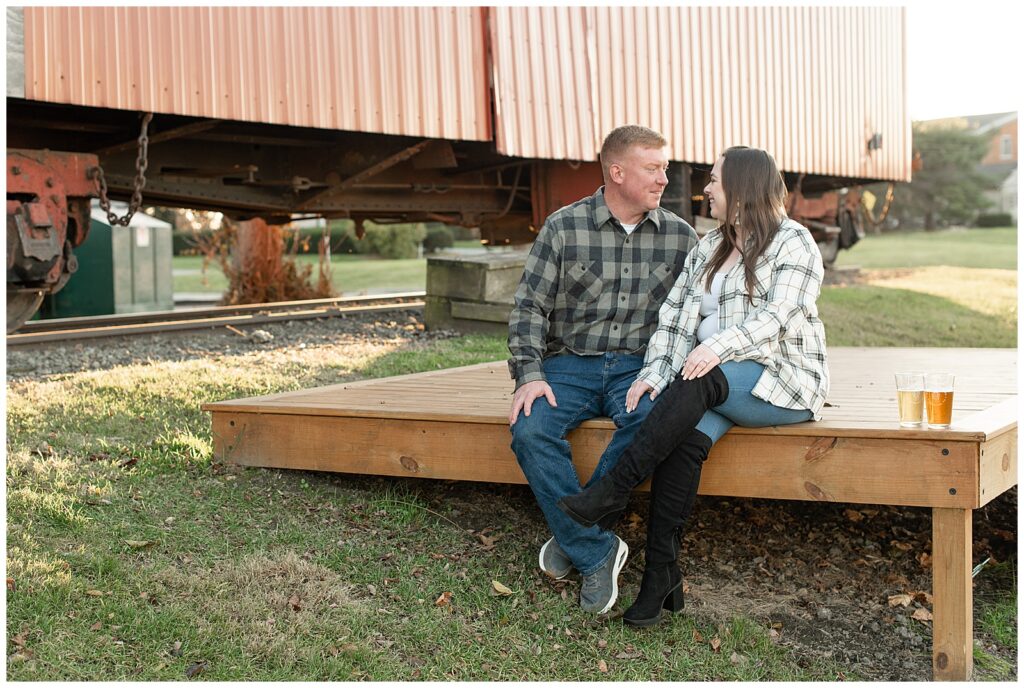 couple sitting on wooden deck by red train as they smile at each other in strasburg pennsyvlania