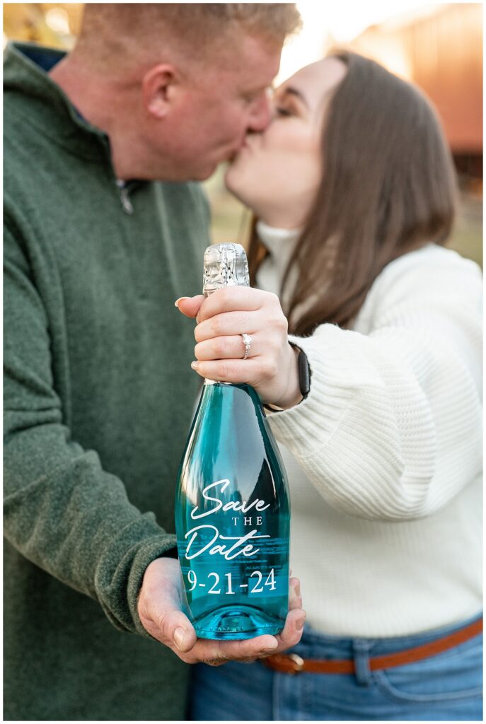 couple kissing and holding out teal bottle with their wedding date on it at bespoke brewery in strasburg pa