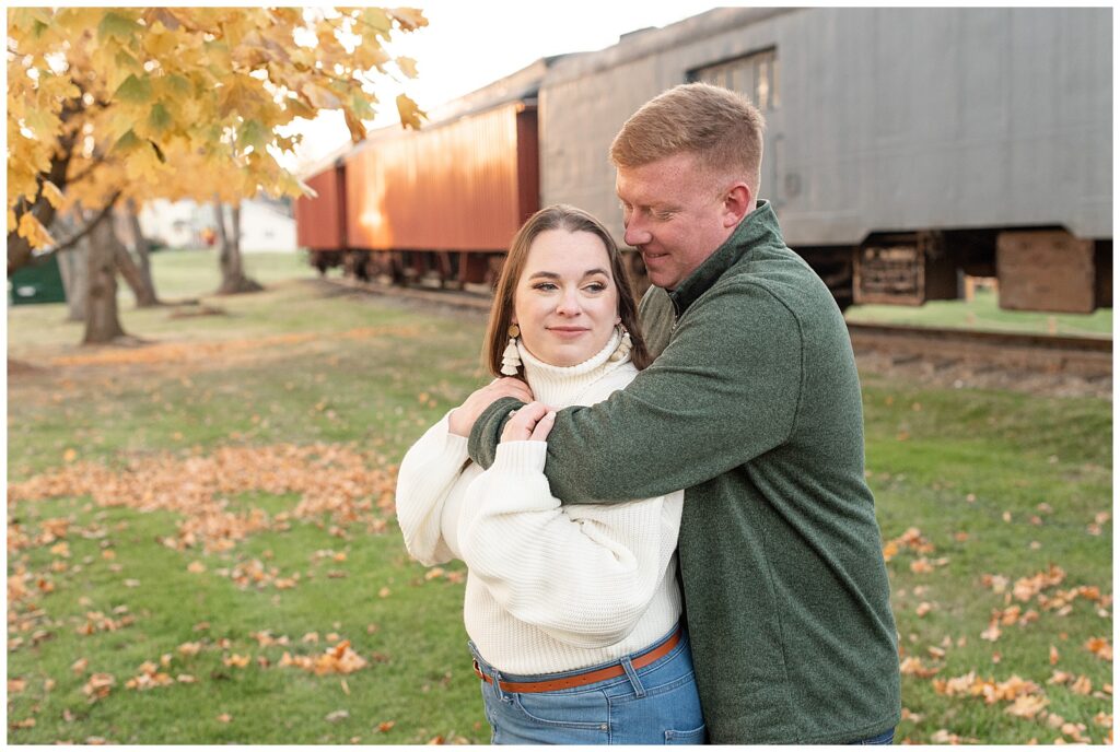 guy hugging girl from behind as she looks off into distance at sunset with colorful fall leaves and train behind them in strasburg pa