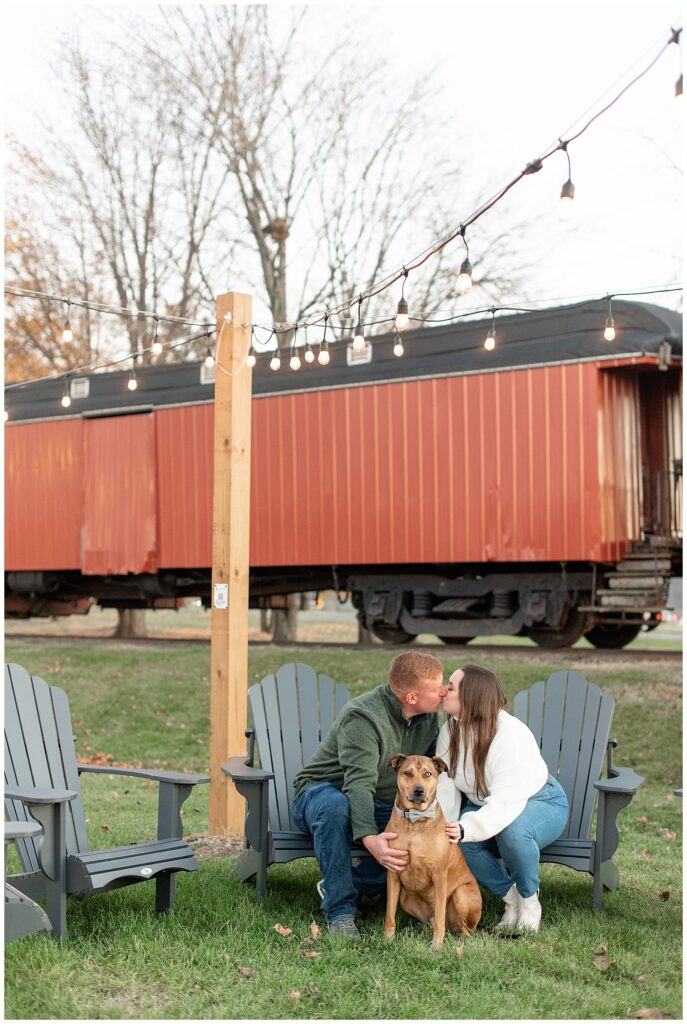 couple sitting in green Adirondack chairs kissing with their pup looking at the camera and red train behind them at bespoke brewery in strasburg pennsyvlania