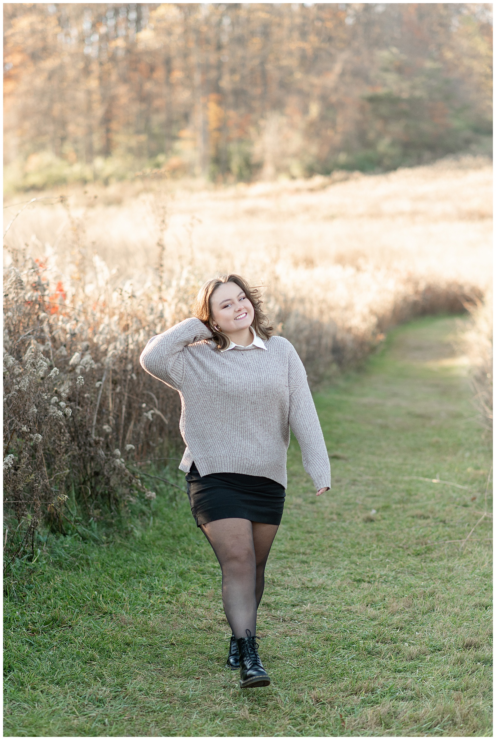 senior girl wearing light gray sweater, black skirt and black tights walking towards camera and putting her hair behind her right ear by tall wild grasses at sunset in stevens, pennsylvania