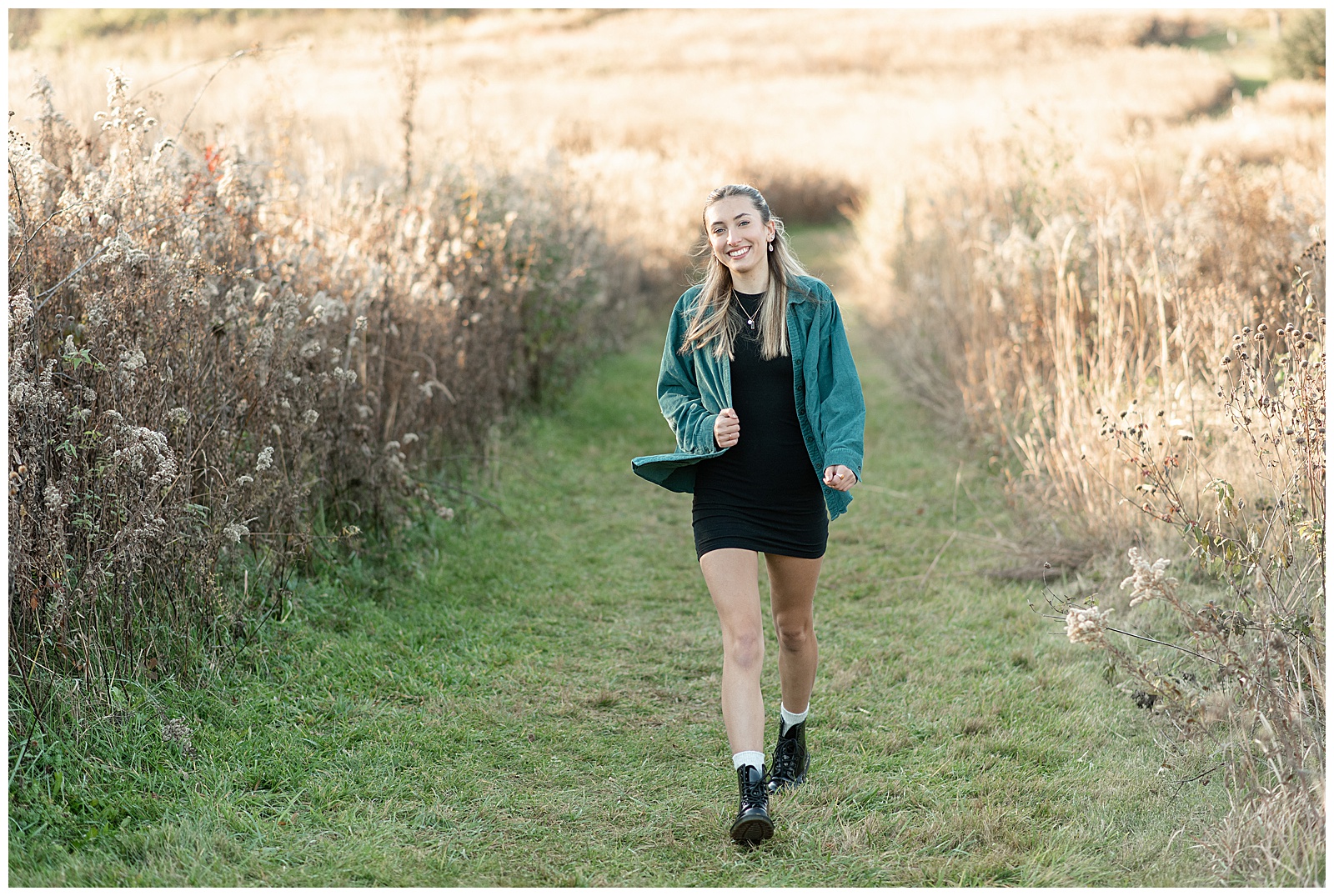 senior girl in black dress with oversized dark green shirt unbuttoned overtop walking in grass path between wild grasses in lancaster county