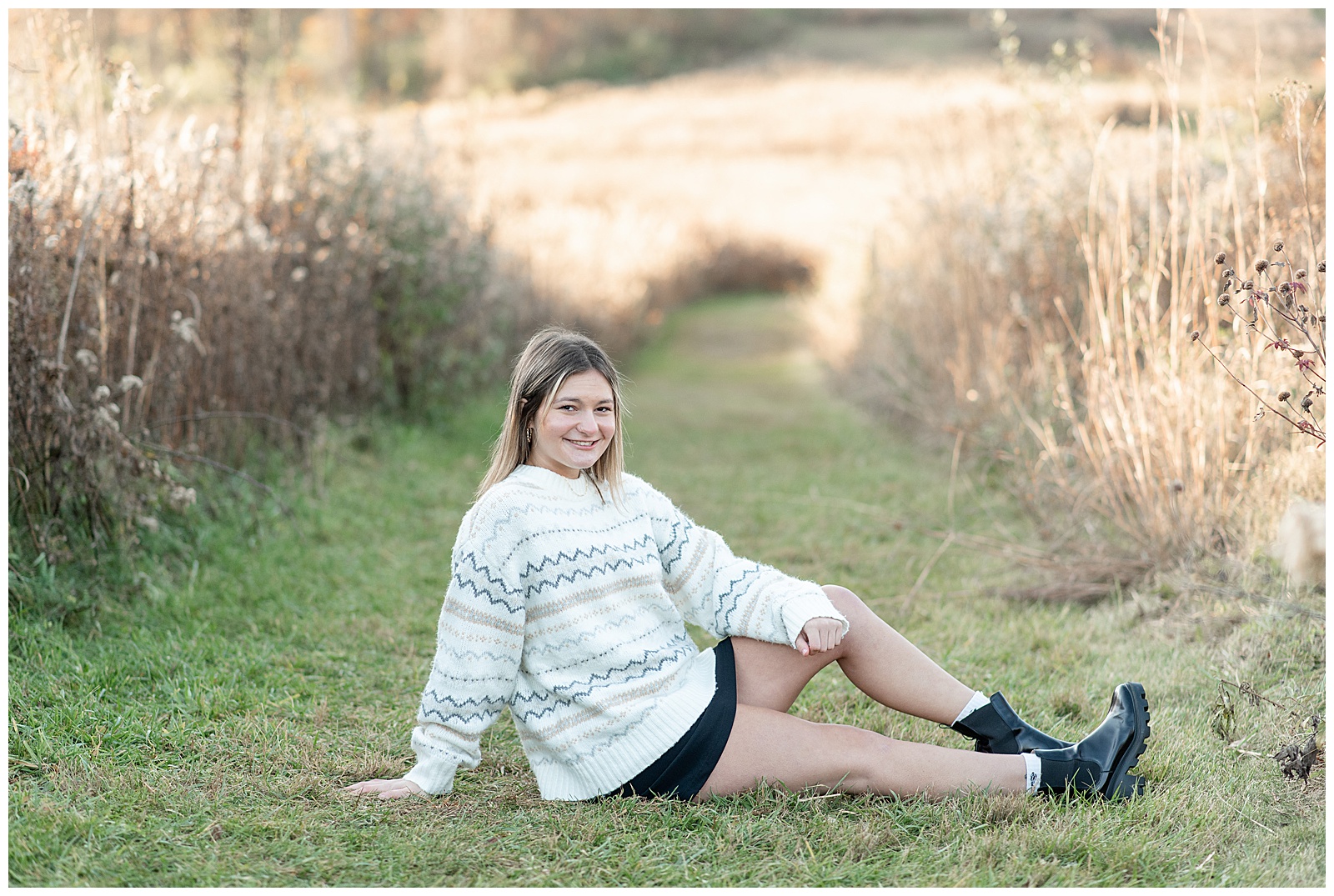 senior girl in white oversized sweater and black mini skirt sitting on grass with legs extended by wild grasses at middle creek