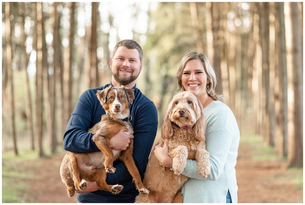 couple each holding a dog smiles at camera by rows of evergreen trees in lancaster pennsylvania