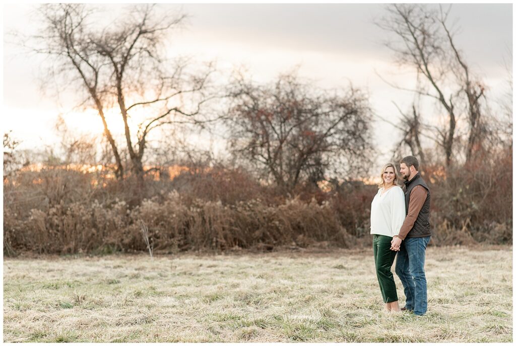 man standing behind woman as she looks at camera and they stand in field at sunset at overlook park