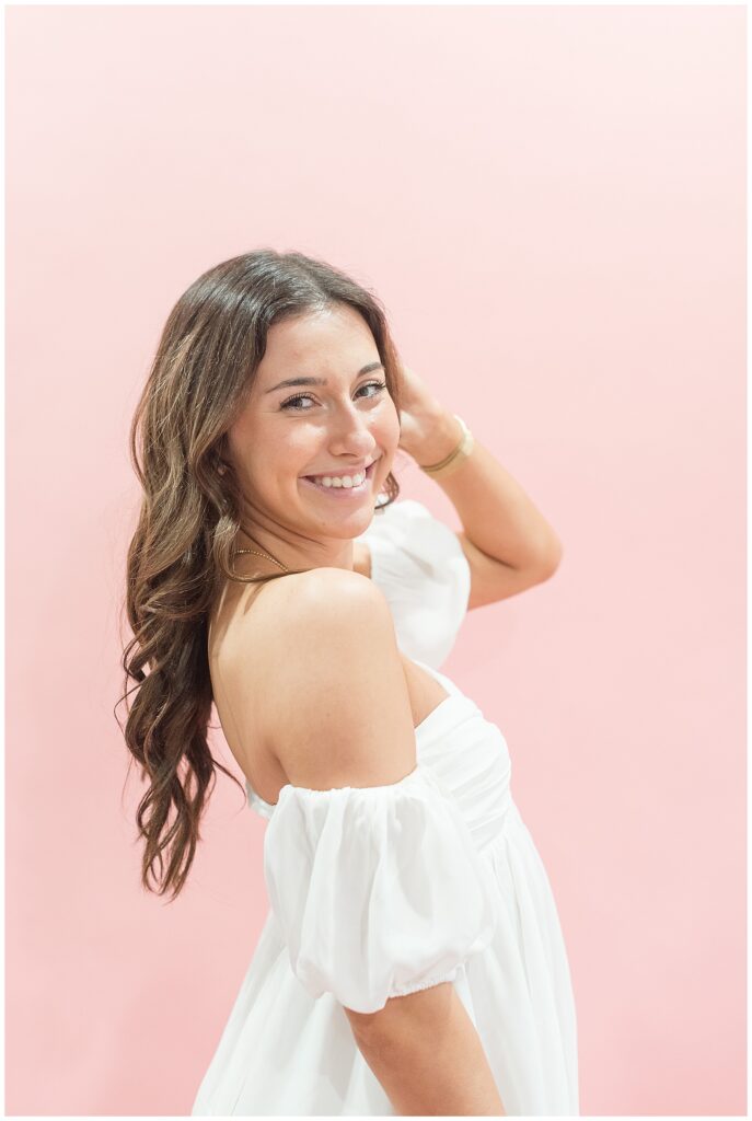 senior girl tucking long brown hair behind her left ear and smiling wearing off-the-shoulder white top at haven studios