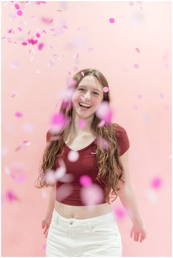 senior girl wear red top and white pants behind sprinkling of pink confetti at haven studios in lancaster pa