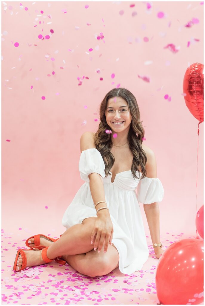senior girl sitting on floor in white dress and red strappy shoes by red balloons at haven studios