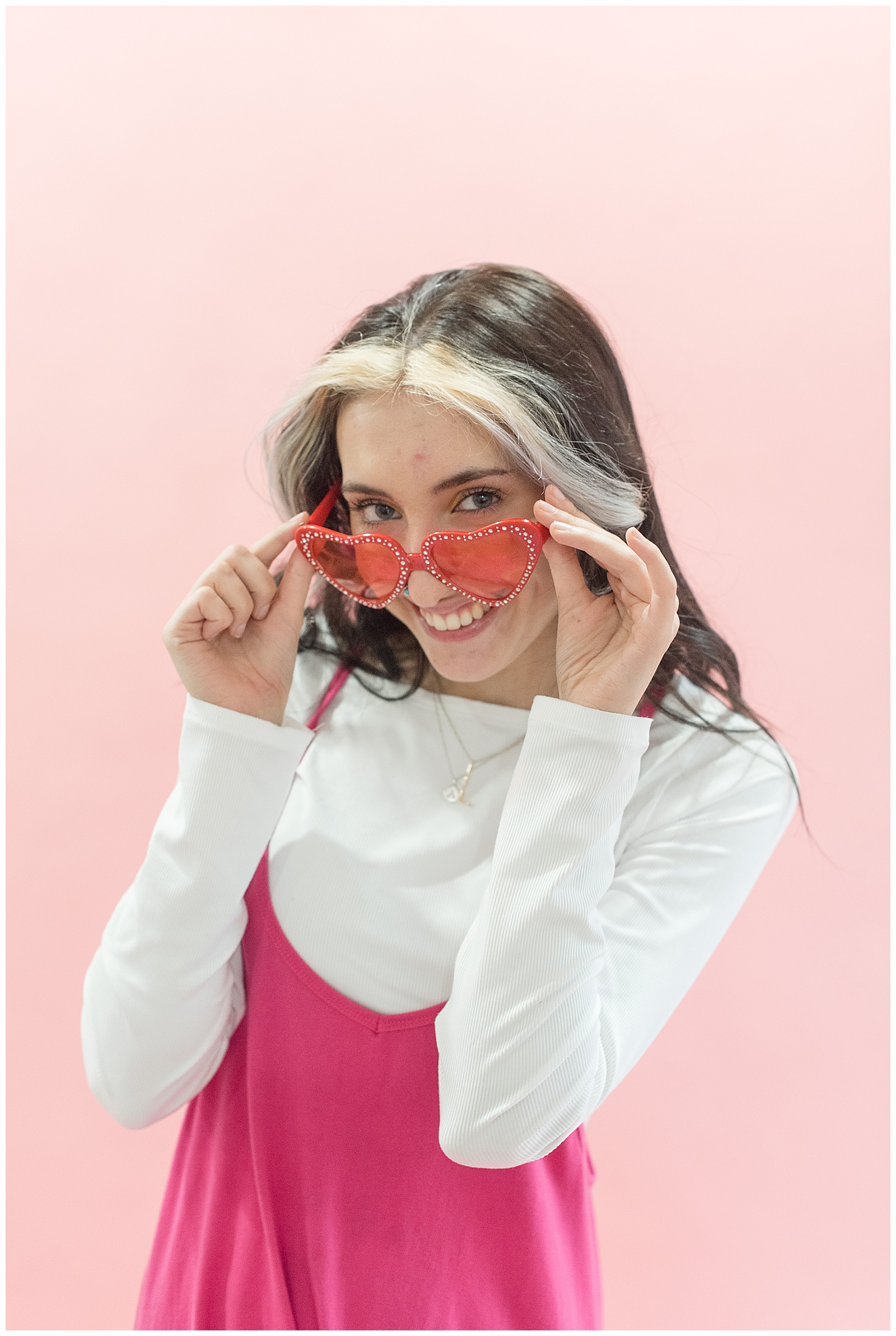senior girl peeking out over the top of her red heart-shaped glasses and smiling at haven studios in millersville pa
