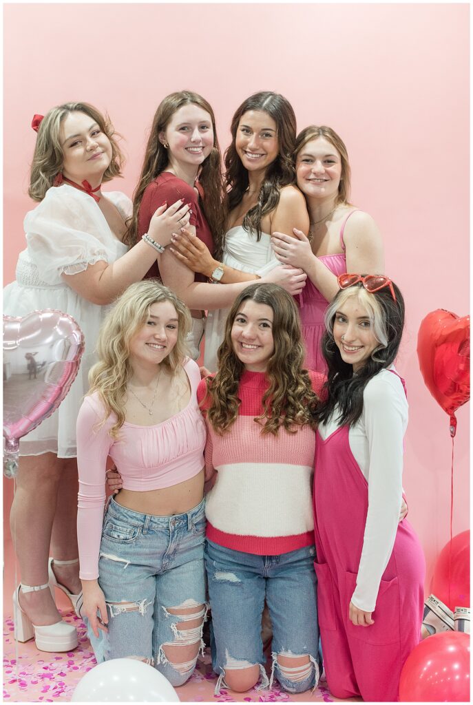 seven senior girls huddled together in shades of red, pink, and white by light pink wall in lancaster county pennsylvania