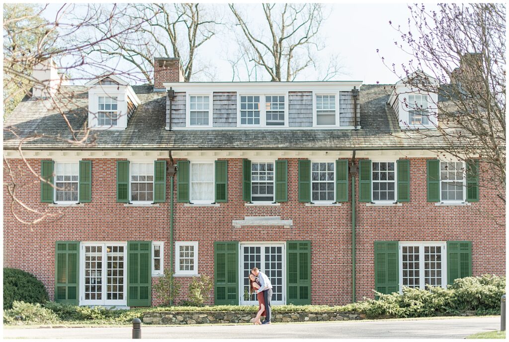 engaged couple hugging outside of red brick home with green shutters at longwood gardens