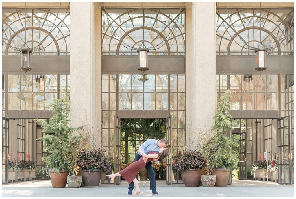 man dips back his bride-to-be as they kiss in front of glass doors at longwood gardens