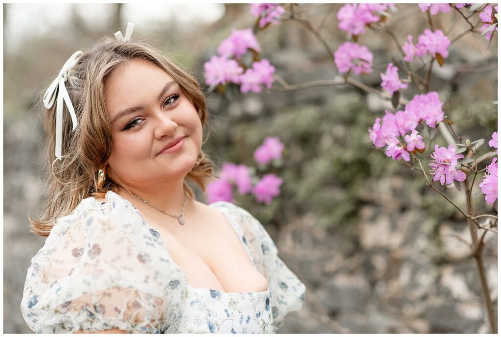 senior spokesmodel in white and blue floral dress smiling at camera by blooming spring bush in chester county, pennsylvania