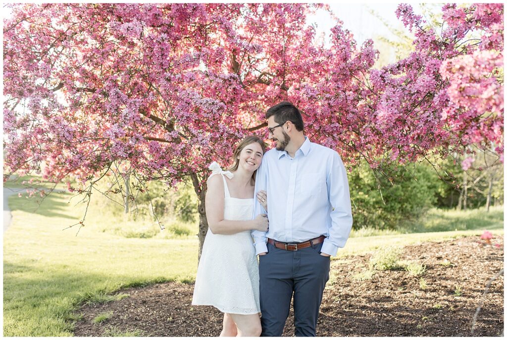 woman in white dress leaning her head against man's right shoulder by blooming pink tree at overlook park