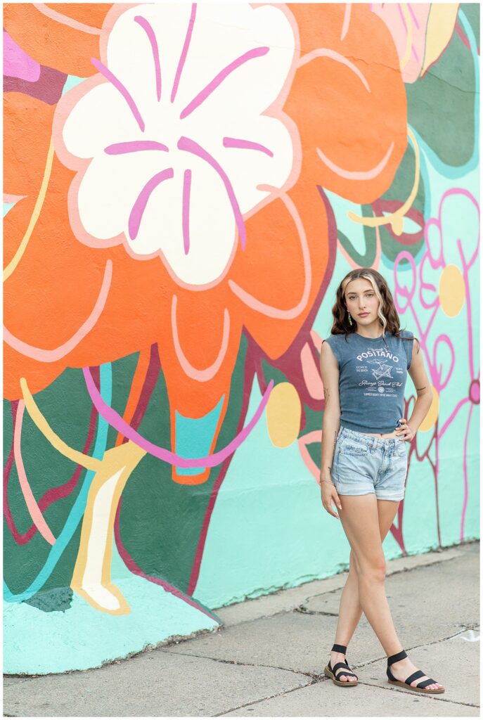 senior girl in blue top and jean shorts with left hand in pocket by colorful flower mural in lancaster county