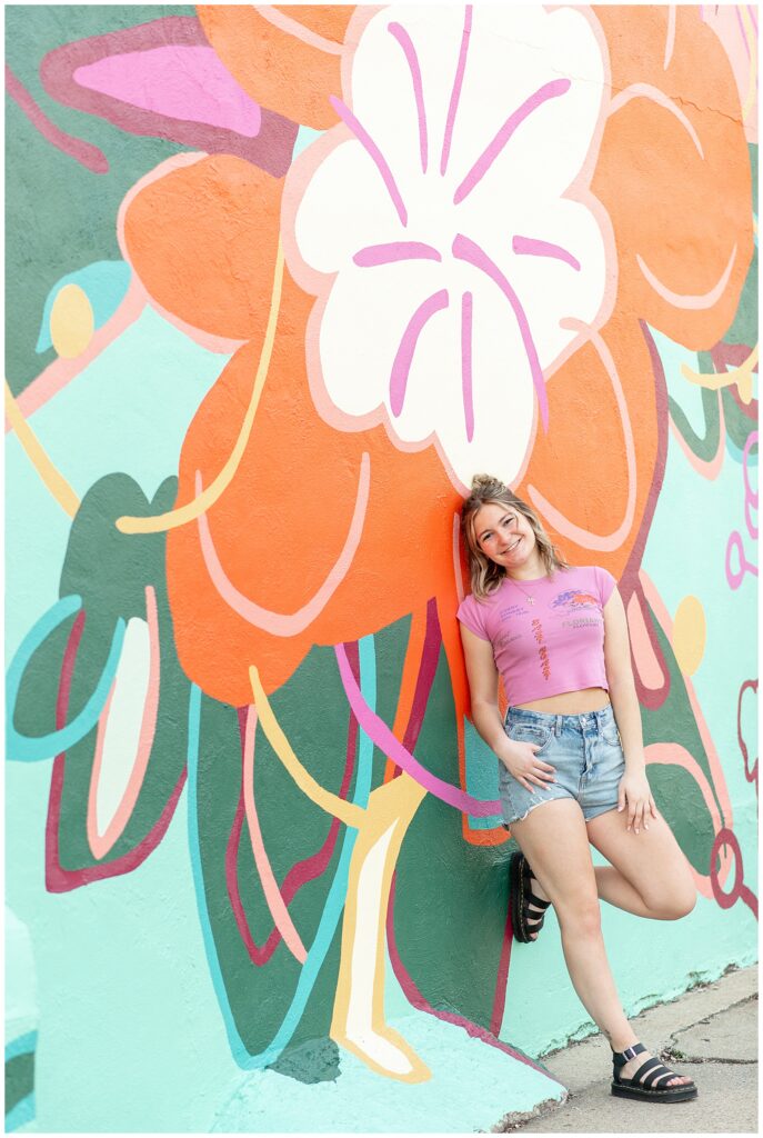 senior girl in pink top and jean shorts leaning against flower mural with left foot bent up against wall in lancaster pa 