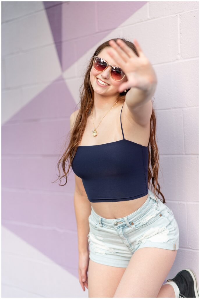 senior girl in navy blue tank top and jean shorts holding her left up in front of her towards camera wearing sunglasses by purple mural in downtown lancaster pa 