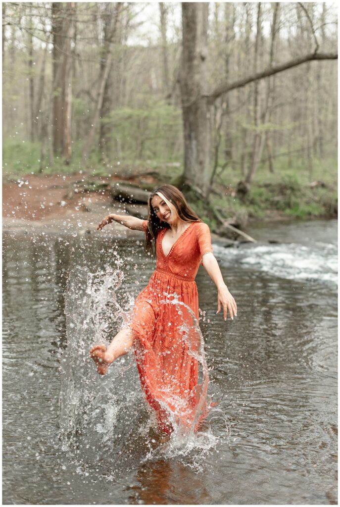 senior spokesmodel in long orange dress standing in stream and kicking the water with her right foot at park in lititz pennsylvania