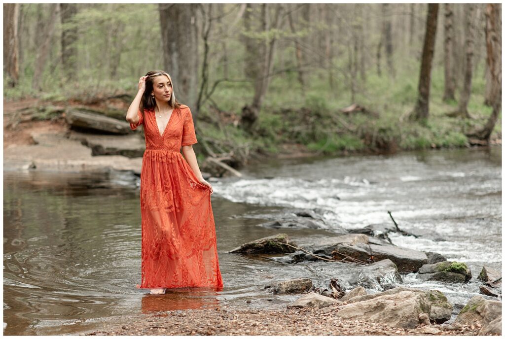 senior girl in long orange dress standing in shallow water and holding edge of dress with her left hand at warwick park