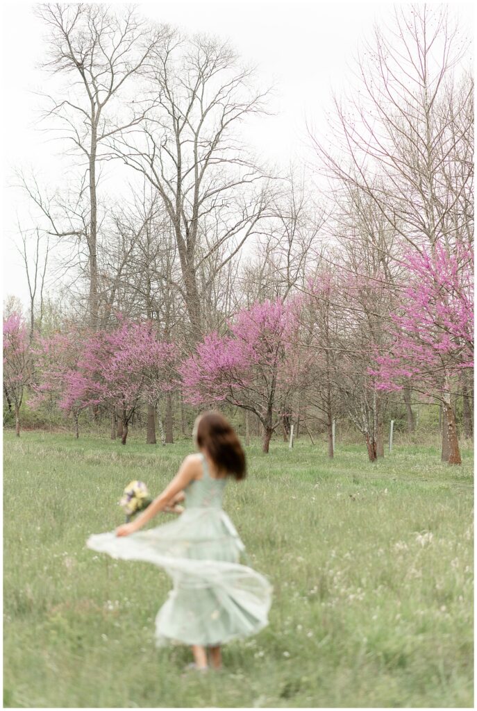 senior girl in long sage green dress spinning holding edge of dress with her back toward camera by blooming pink trees at warwick park