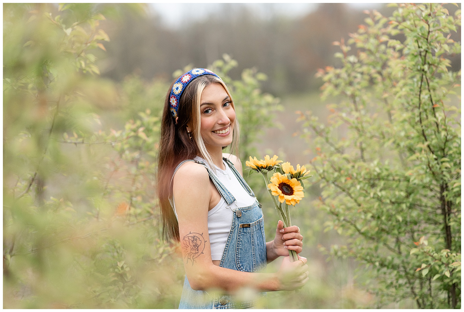 senior girl in denim overalls and white tank top holding bouquet of sunflowers and standing in field in lancaster pa