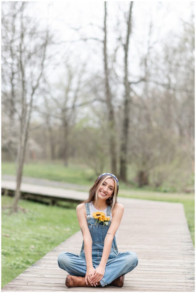 senior girl sitting on wooden walkway as she smiles at camera at park in lititz pennsylvania