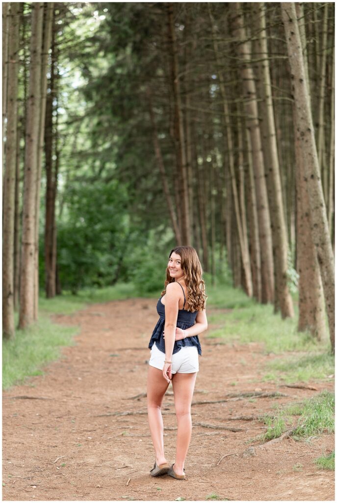 senior girl walking away from camera and looking back over left shoulder by row of evergreens at overlook park