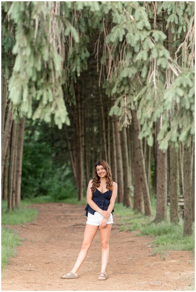 senior girl in black tank top and white shorts by row of trees at overlook park in lancaster county