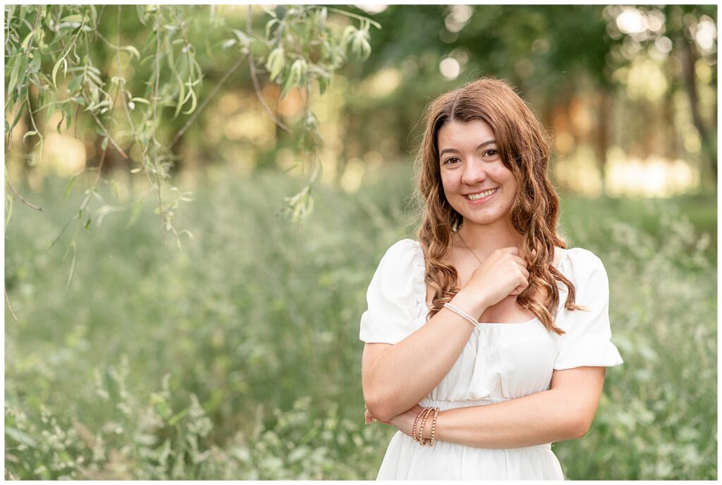 senior girl in cute white dress standing by tall grasses with arms folded against her chest in lancaster pa