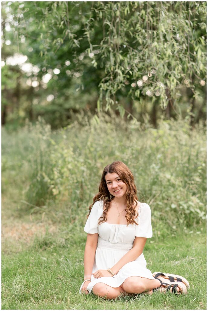 senior girl sitting in grass with legs folded under her by tall grasses at overlook park