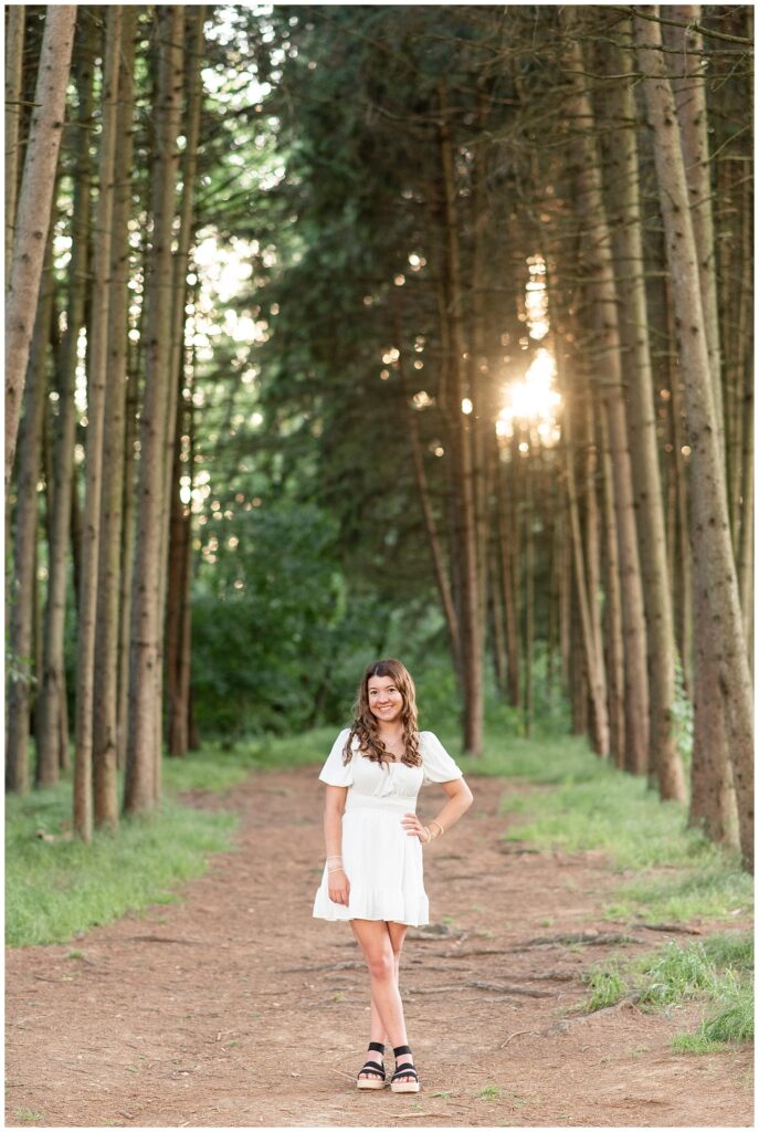 senior girl in white dress by rows of tall evergreen greens at sunset at overlook park