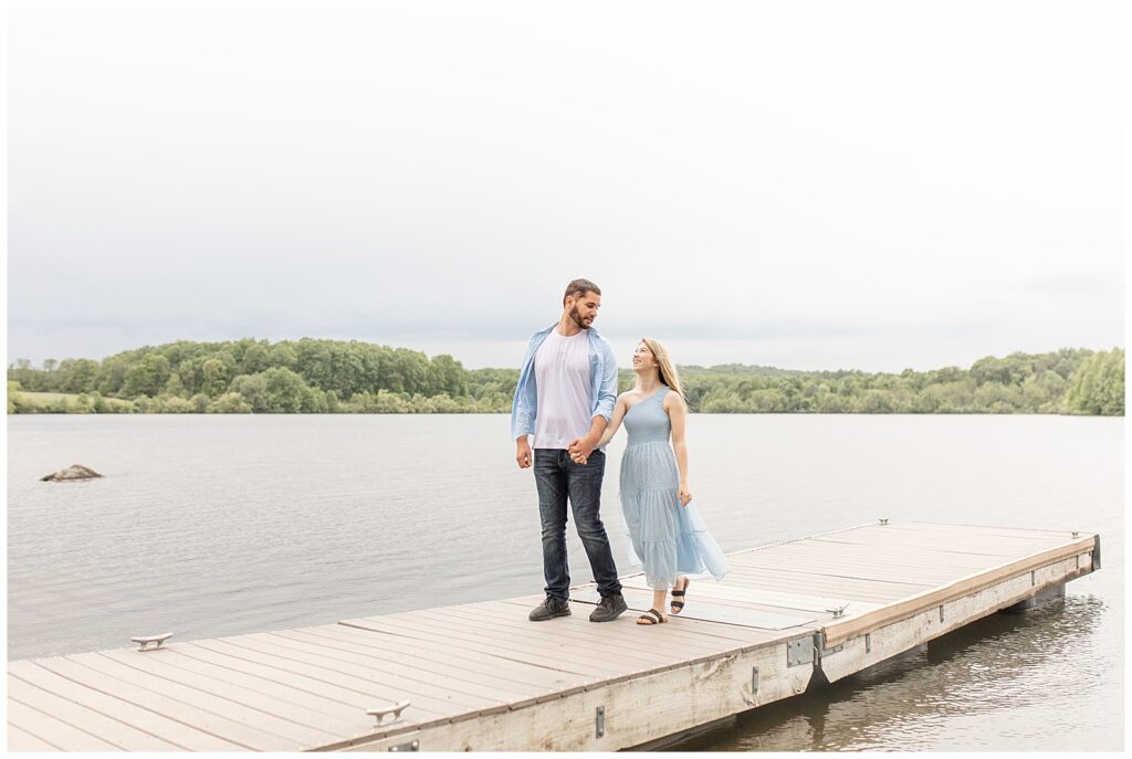 engaged couple holding hands and walking along wooden dock of pond at hibernia park