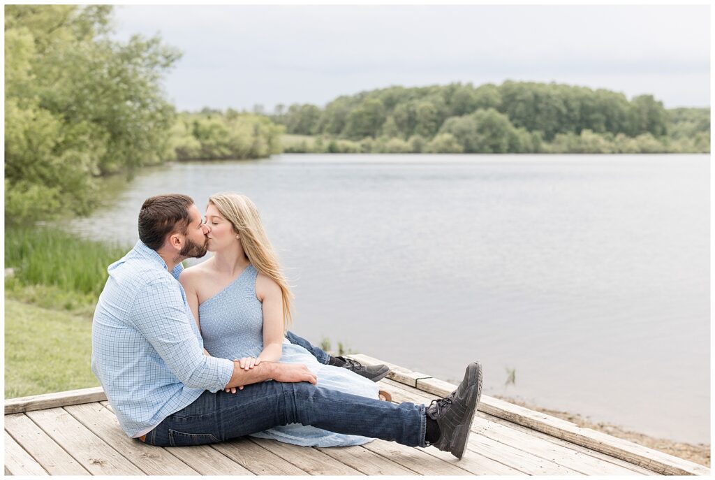 engaged couple sitting on wooden dock and kissing by pond at hibernia park in chester county