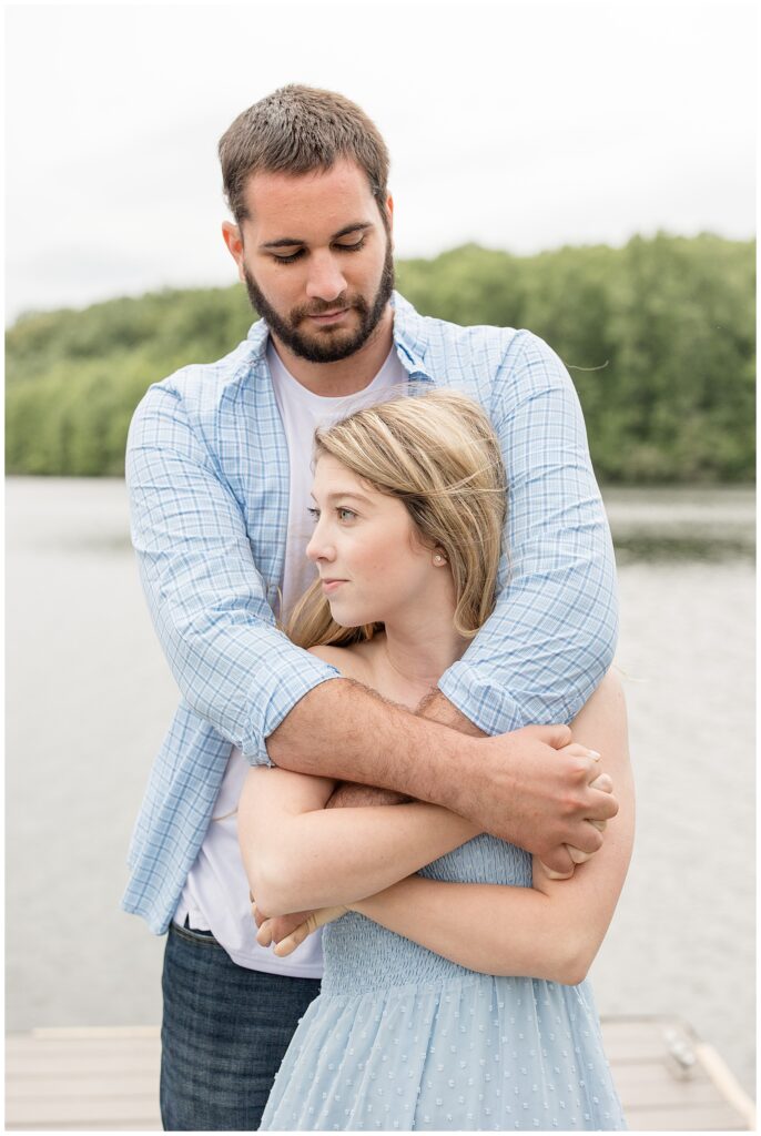 man hugging woman from behind as he looks down and she looks to the right by pond at hibernia park