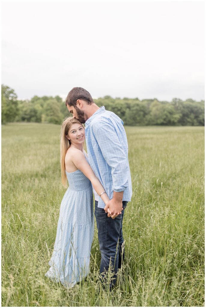 engaged couple hugging and holding hands as man kisses top of woman's head in grass field at hibernia park