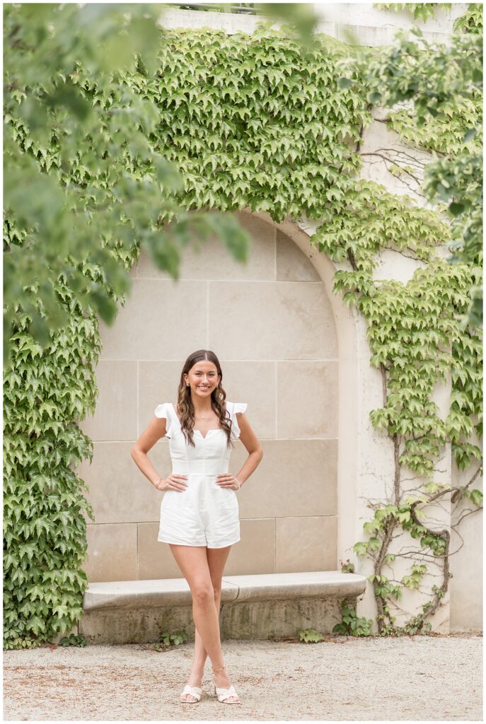 senior girl in cute white romper by ivy archway with her hands on her hips at longwood gardens