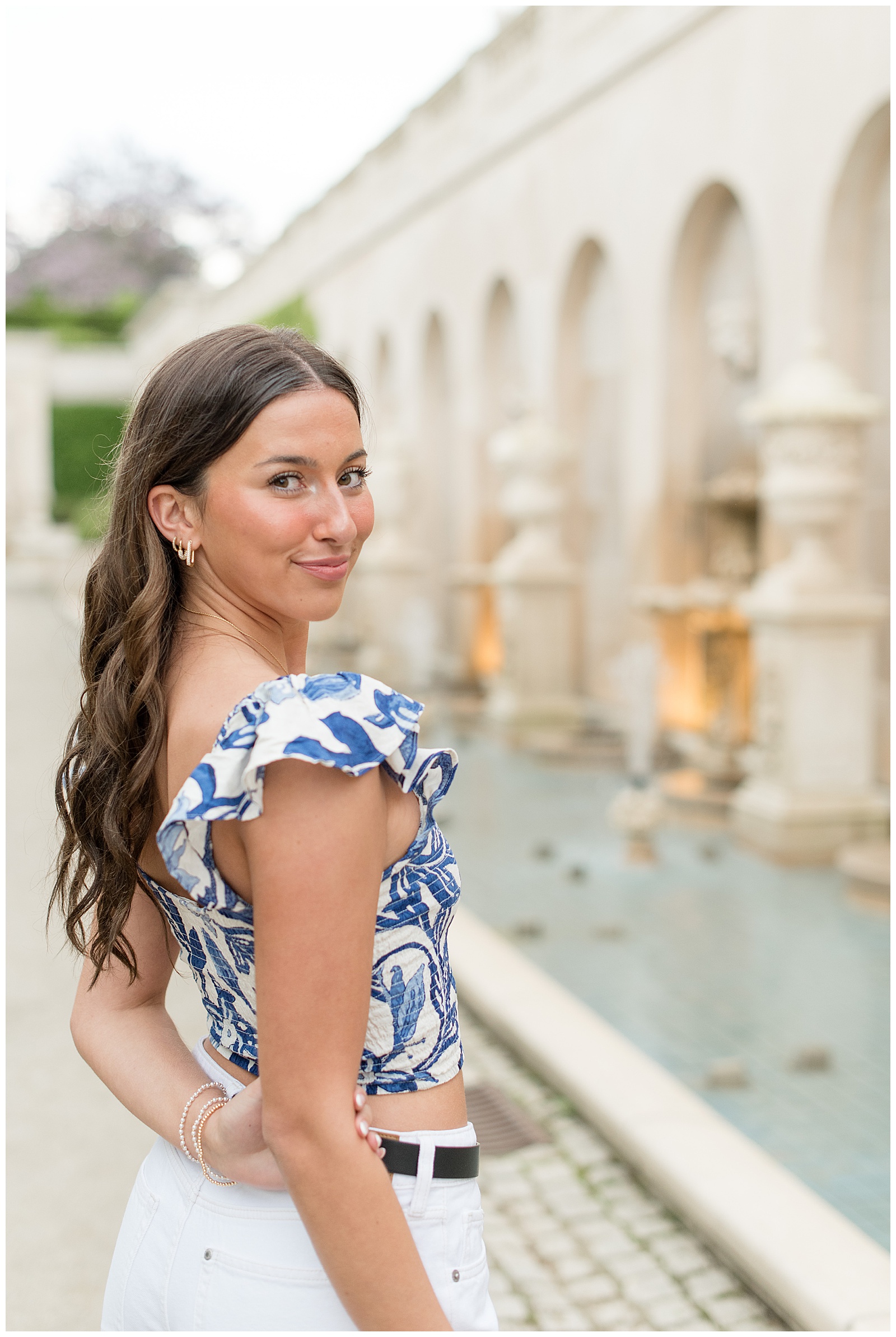senior girl in blue and white top with white jeans looking over her right shoulder by fountain at longwood gardens