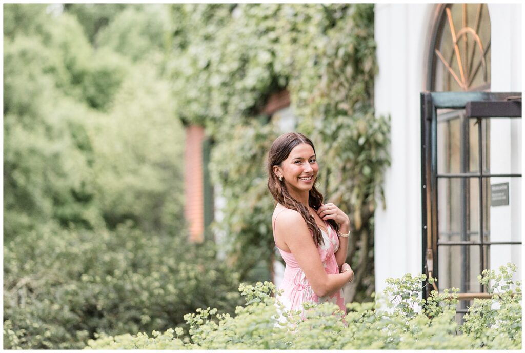 senior girl in pink and white flowy dress looking over right shoulder by ivy and greenery at longwood gardens on overcast spring evening