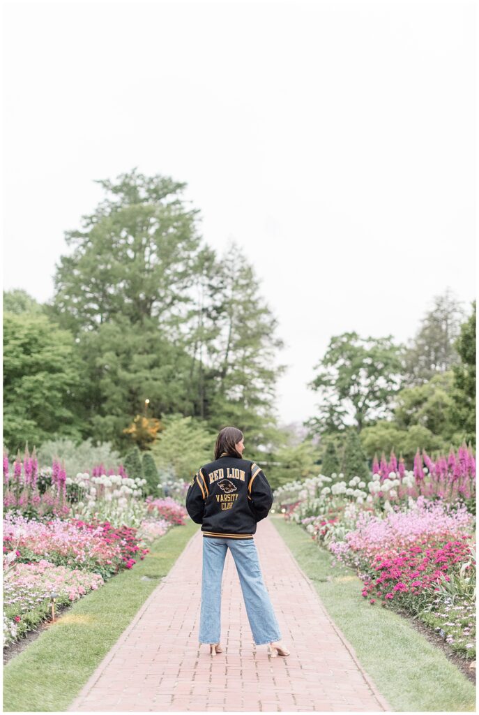 senior girl in red lion black and yellow varsity jacket and blue jeans standing on brick walkway with her back to the camera at longwood gardens